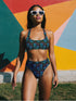 Two-piece high-waisted bikini set featuring an all-over print of blue skulls with purple bows and multicolored stars on a dark background. Includes a deep V-neck bikini top with wide straps and matching high-rise bottoms. Made from soft, stretchy fabric with a smooth black inner lining. The design combines bold, edgy graphics with a flattering retro-inspired silhouette.